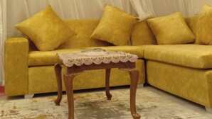 A yellow sofa with matching cushions, a small wooden side table with a lace tablecloth, and a white curtain in the background. - 2Bed Sea View Apartment - Sleeps 5 w Free Parking (Sharm El-Sheikh)