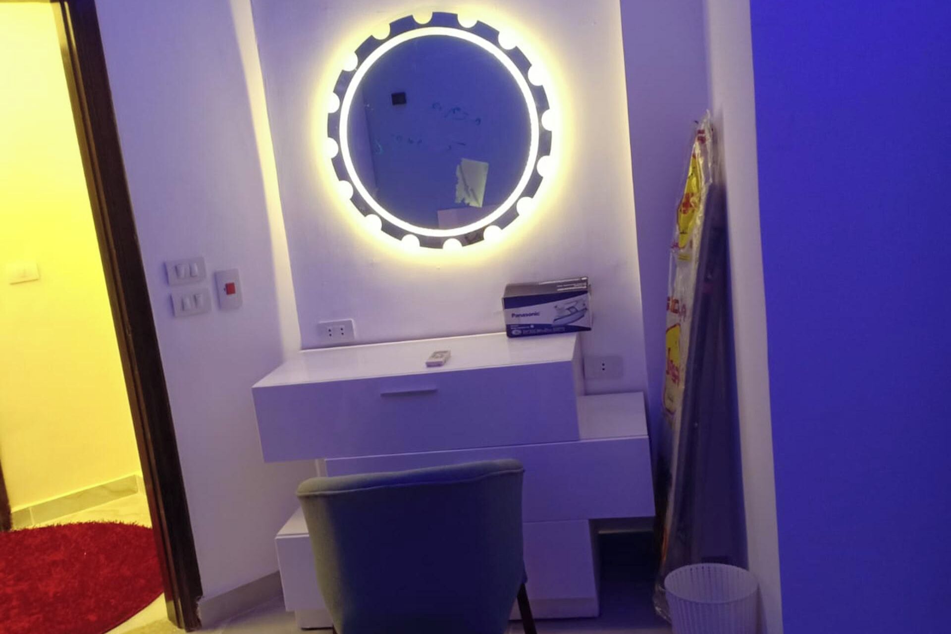 A bathroom with a vanity, a mirror with lights, a towel rack, and a trash bin.