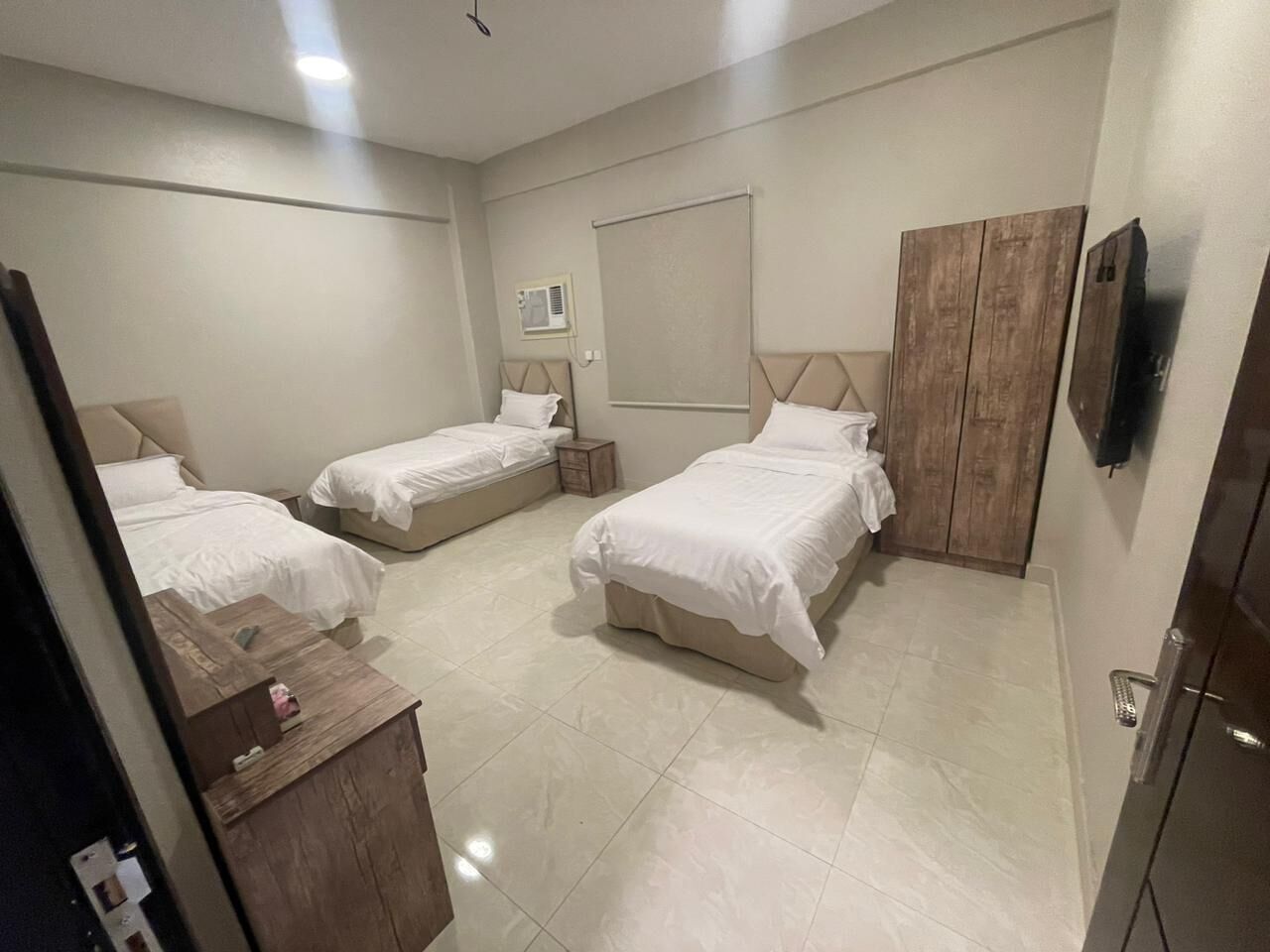 A hotel room with three beds, a wooden wardrobe, a small bedside table, a TV mounted on the wall, and a window with an air conditioning unit.