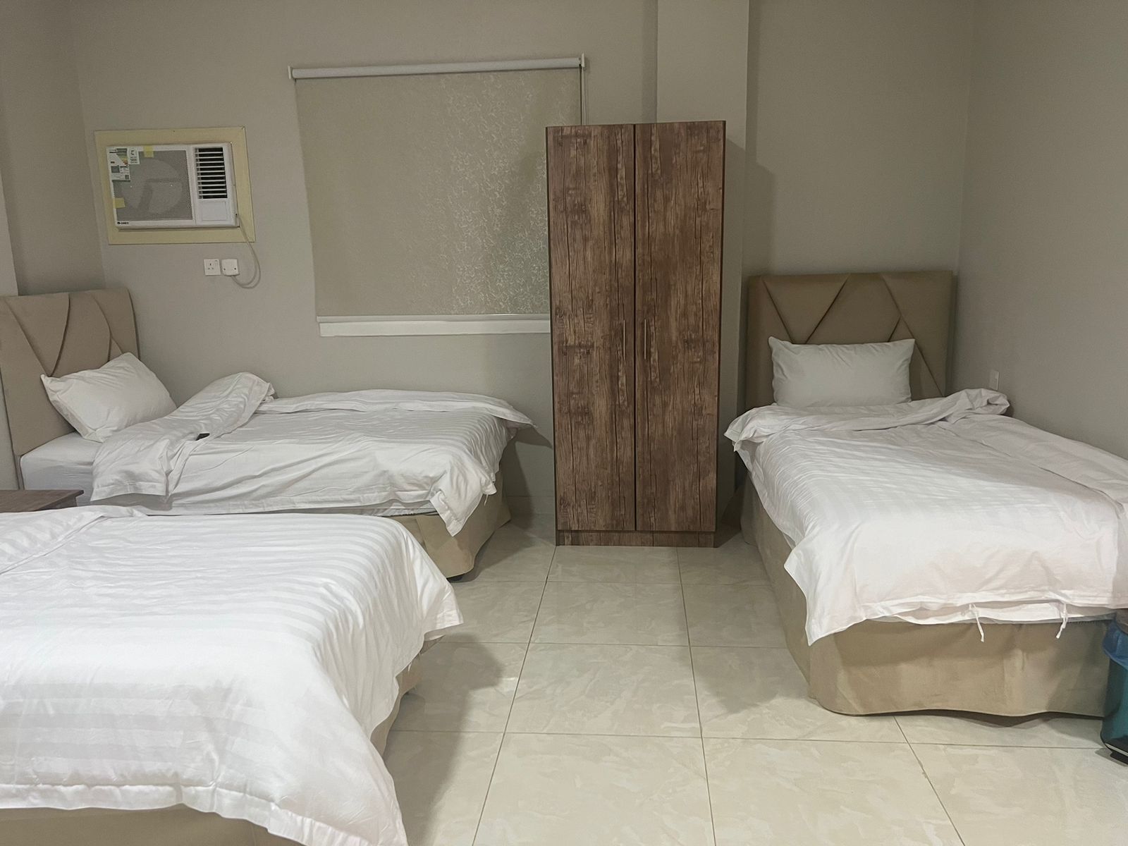 A bedroom with two single beds, a wooden wardrobe, a window with blinds, and an air conditioning unit on the wall.
