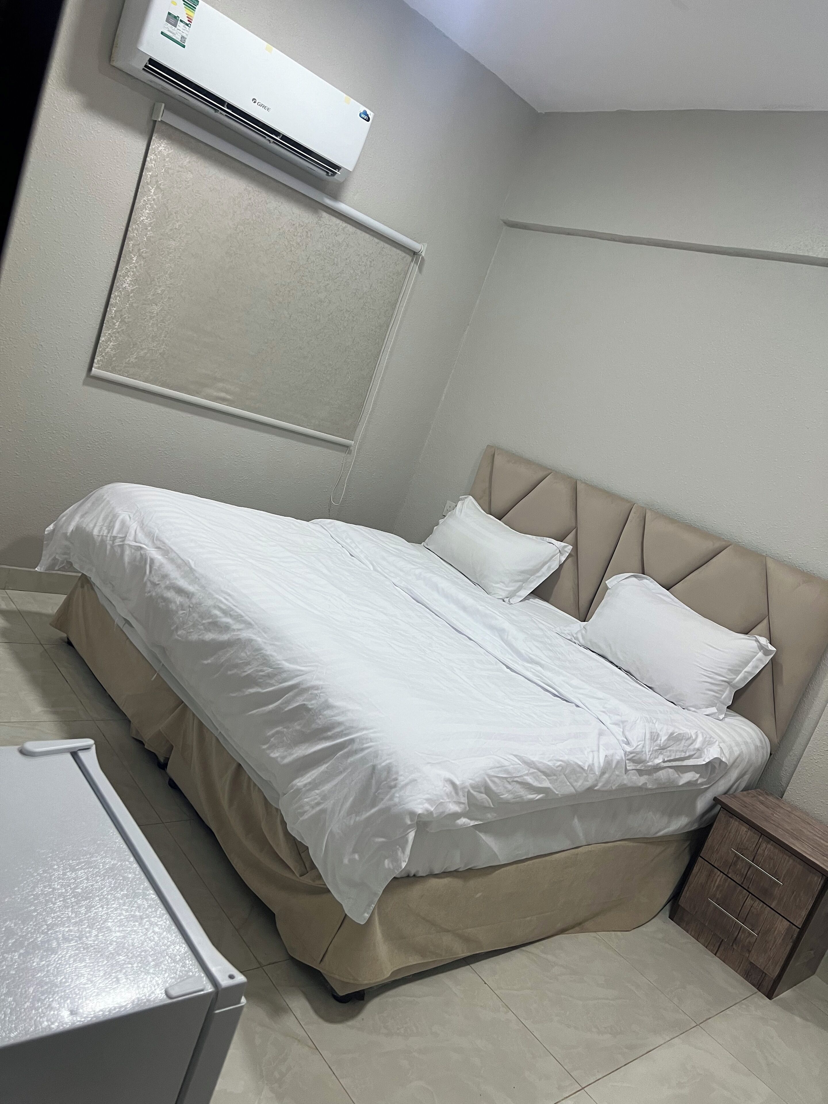 Een net opgemaakt bed met witte lakens, een beige hoofdbord en een klein houten nachtkastje met lades.