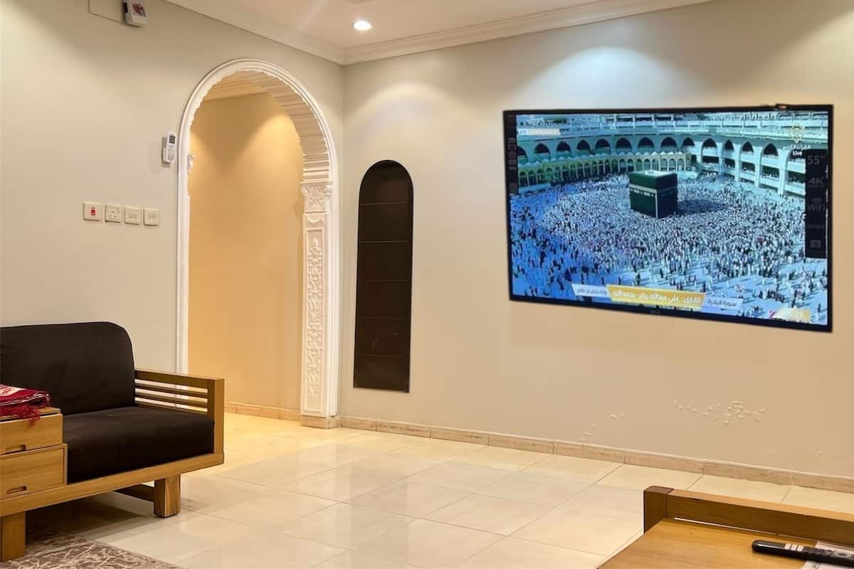 A modern hotel room with a large TV displaying a crowd at a significant religious site, a black sofa, a small wooden table, and a lamp.