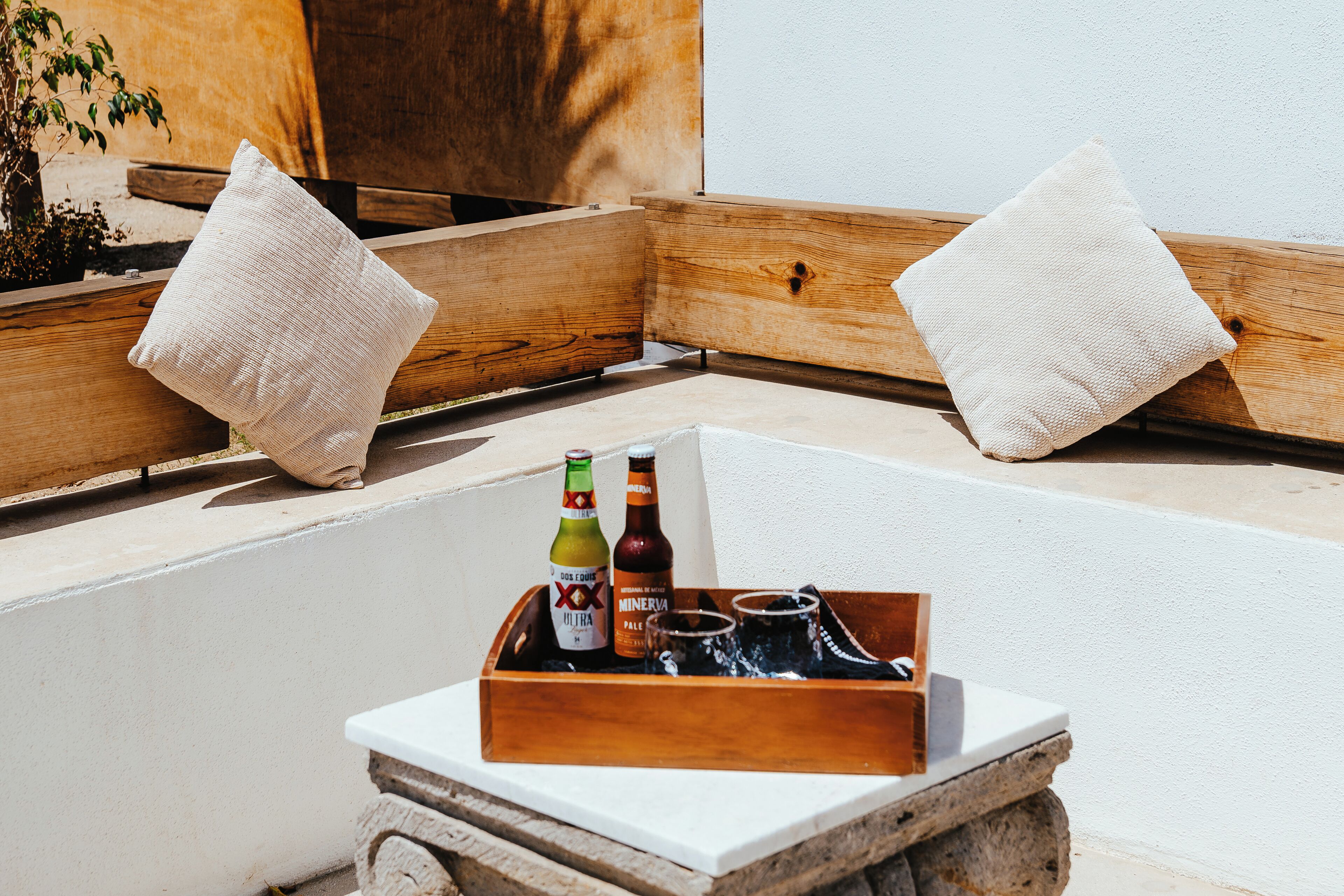 A wooden bench with cushions, a tray with bottles and glasses, and a small plant.