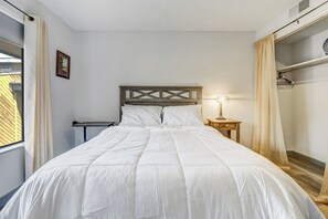 A neatly made bed with a wooden headboard, a bedside table with a lamp, and a window with curtains. - Trailhead Terrace (Colorado Springs)