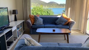A living room with a sofa, a coffee table, and a TV. There is a bookshelf with books and a remote control. The room has a large window with a view of a lake and hills.