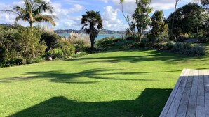 A well-maintained lawn with a wooden deck, surrounded by various tropical plants and trees, with a view of a body of water and hills in the background.