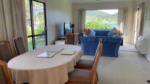 A room with a dining table, chairs, a sofa, and a view of greenery and mountains through large windows.