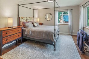 Un lit à baldaquin avec une tête de lit en bois, une table de chevet avec des tiroirs, un miroir rond accroché au mur et une fenêtre avec des rideaux.