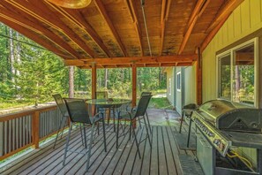 Une terrasse couverte, dotée d’un barbecue et de chaises, entourée d’une rambarde en bois et offrant une vue sur une forêt.
