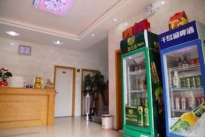 Zona de recepción de un hotel con un mostrador de recepción, una máquina expendedora verde y un refrigerador azul lleno de bebidas.