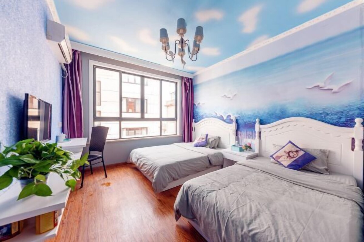 A room with two beds, a desk, a chair, and a large window.