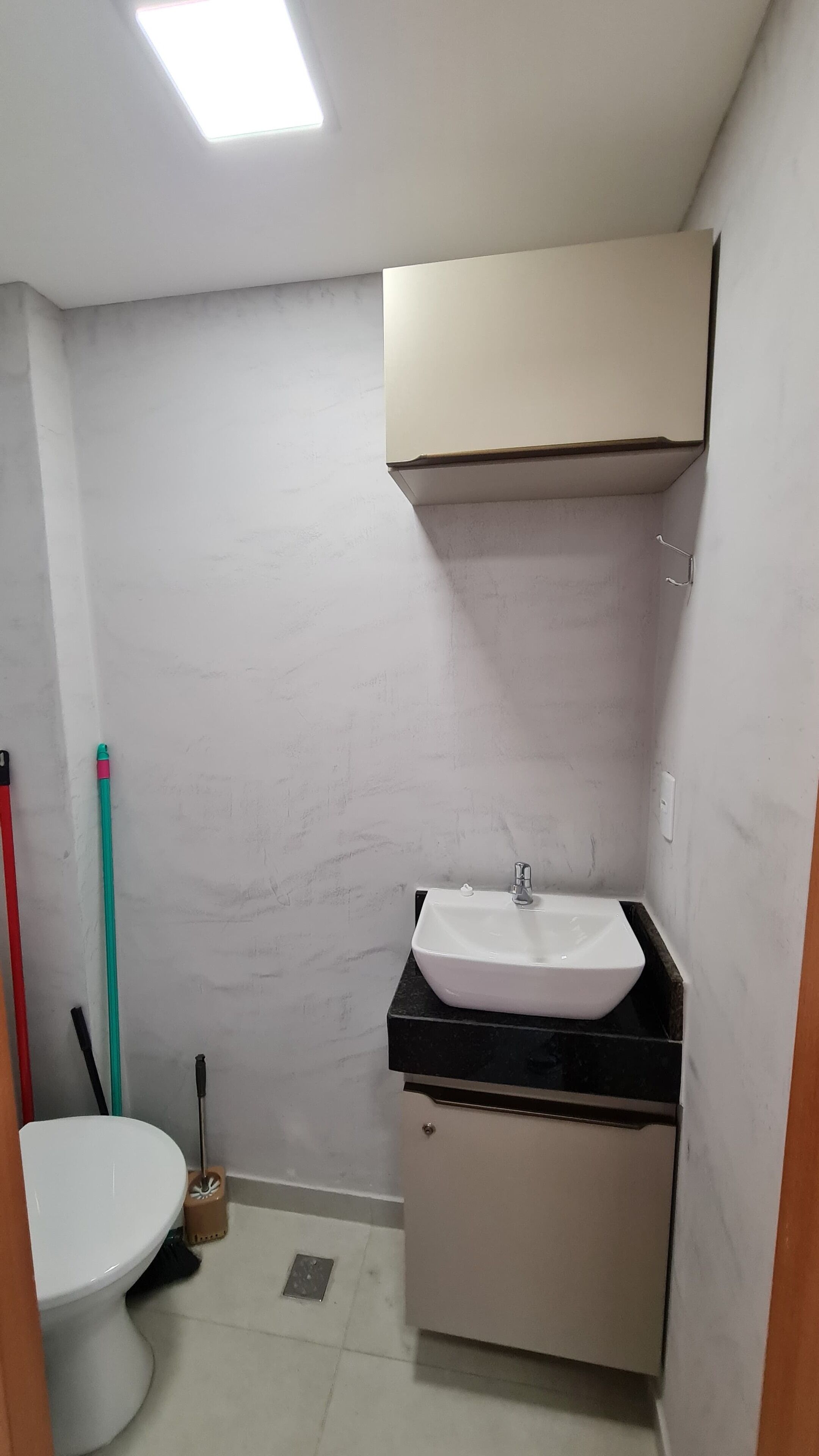 Un baño pequeño con un lavamanos blanco, un espejo y un inodoro. Hay utensilios de limpieza en el suelo.