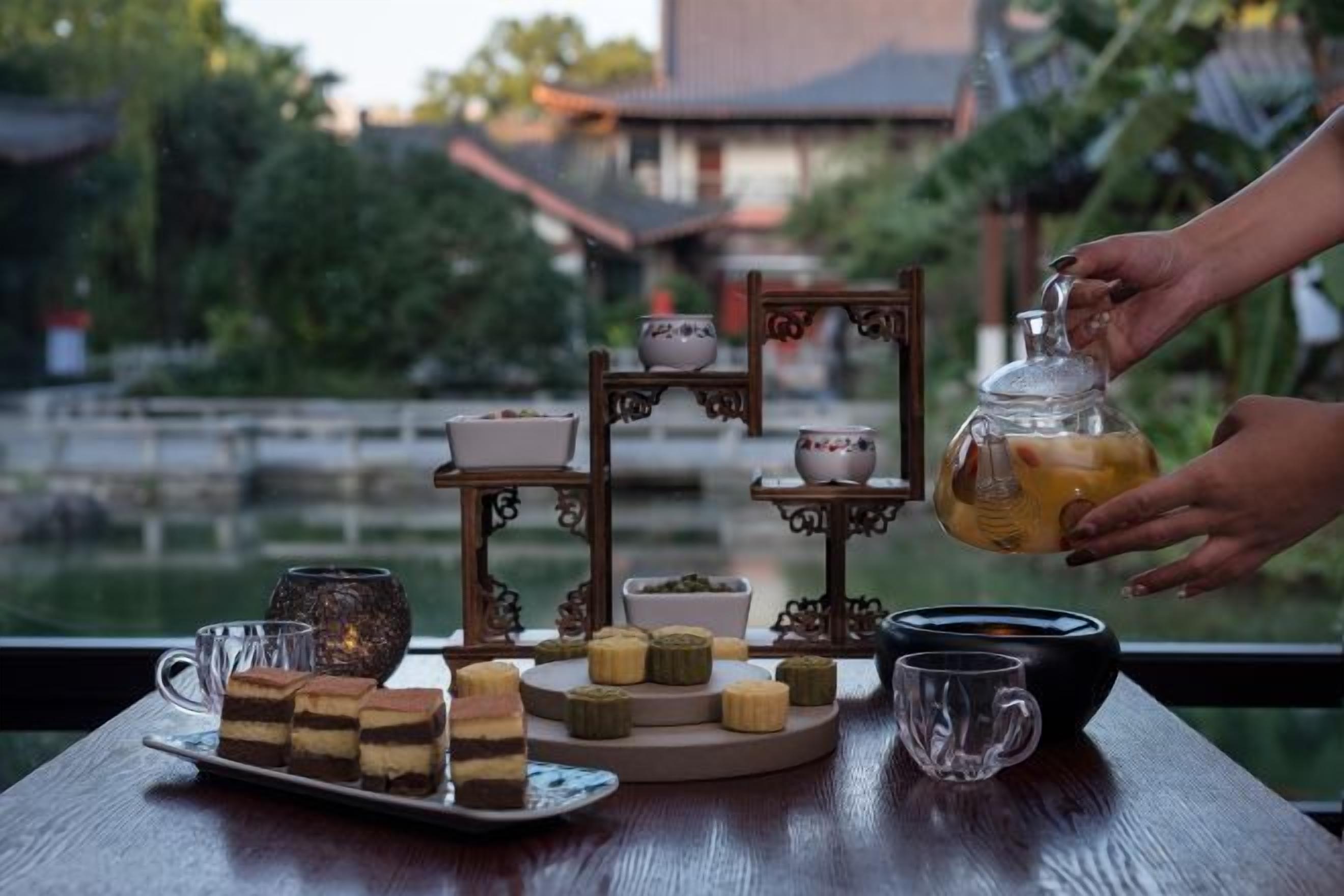 Une table dressée avec divers aliments et une théière, le tout mis en valeur par une vue sur un bâtiment traditionnel et une végétation luxuriante.