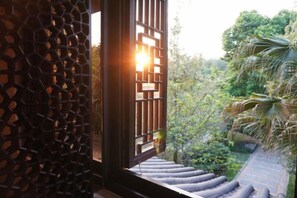 Una ventana con un diseño geométrico, vista a un jardín con árboles y una escalera que lleva a una azotea.