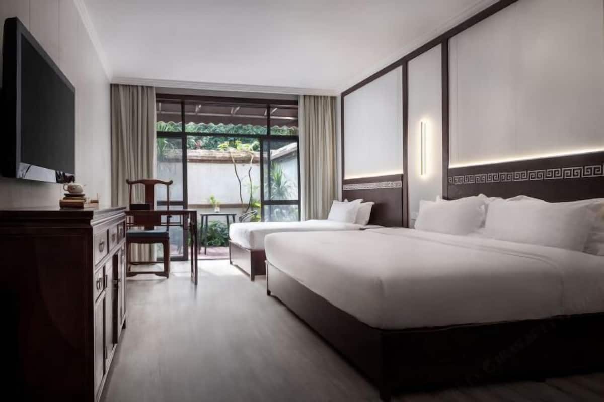 A hotel room with a large bed, a TV, a desk with a chair, and a view of greenery through the window.