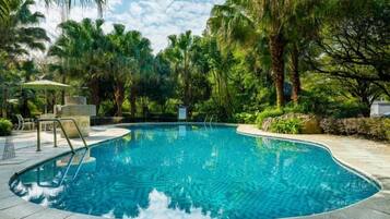 Una piscina rodeada de palmeras y follaje, con zona de estar y un espacio de descanso cubierto.