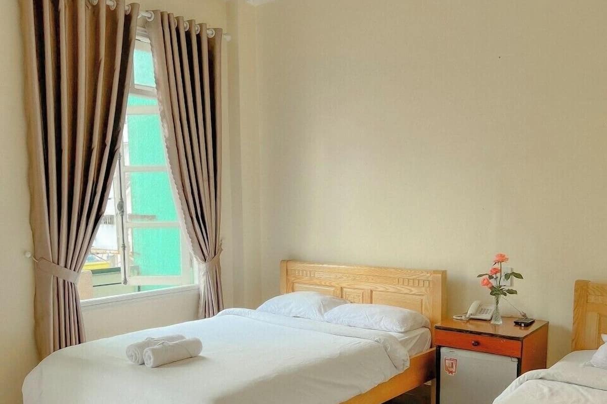 A hotel room with two beds, a small bedside table with a phone and a vase of flowers, and a window with curtains.