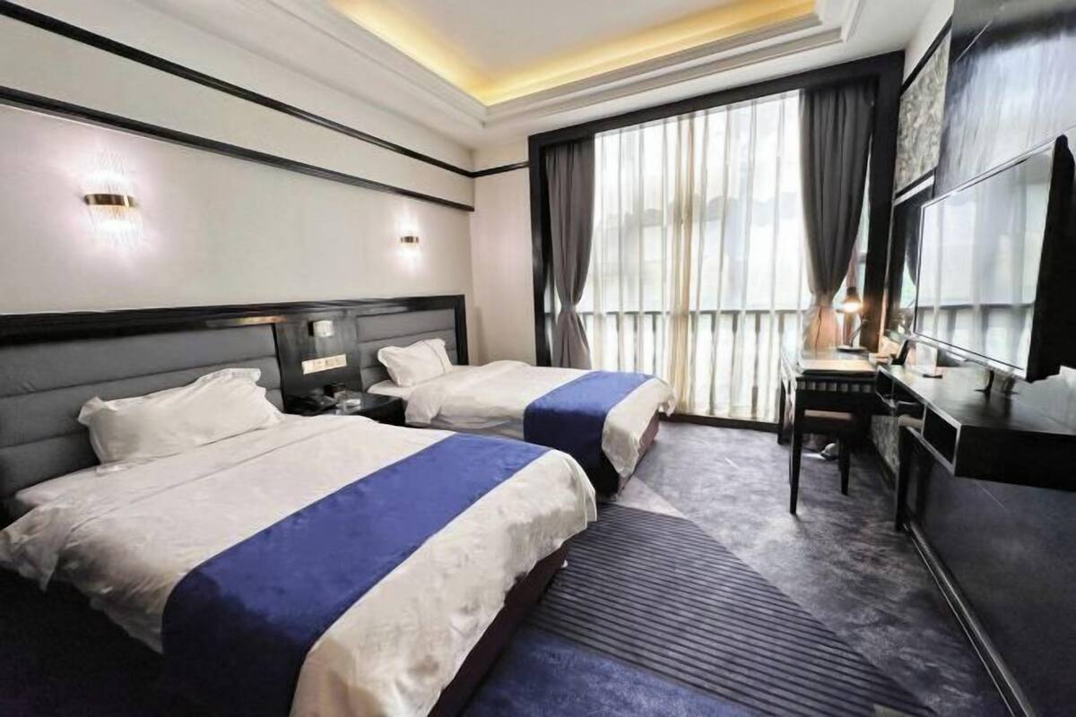 A hotel room with two beds, a desk, a chair, a TV, and a large window with curtains.