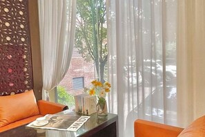 A room with a sofa, a table with a vase of flowers, and a view through a window with curtains. - TS Business Hotel (Meishan)
