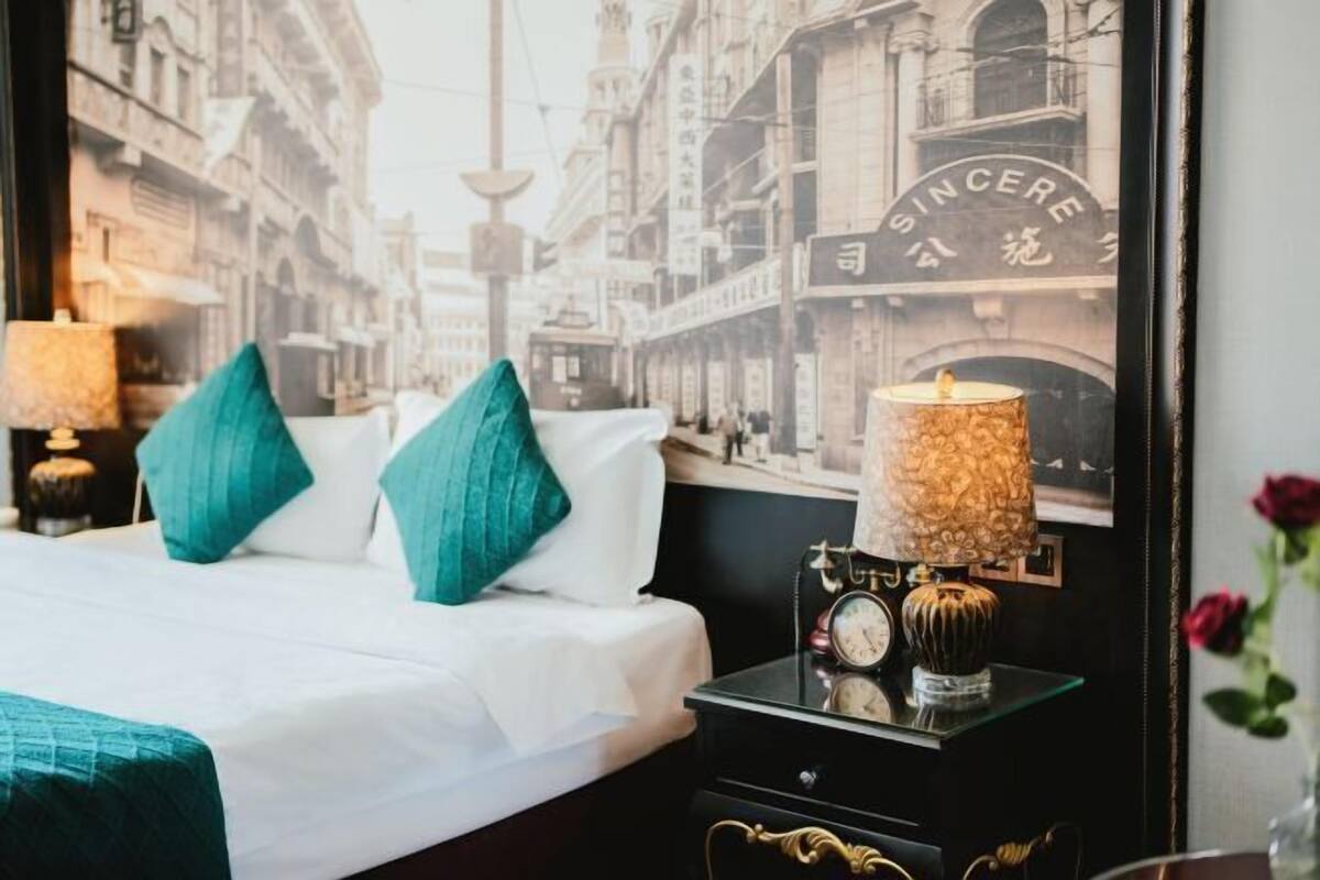 A bed with teal pillows, a nightstand with a clock and a lamp, and a wall with a cityscape and 'SINCERE' sign.