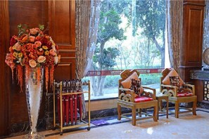 A room with a large floral arrangement, two wooden chairs with cushions, a window with a view of trees, and a wooden cabinet. - TS Business Hotel (Meishan)