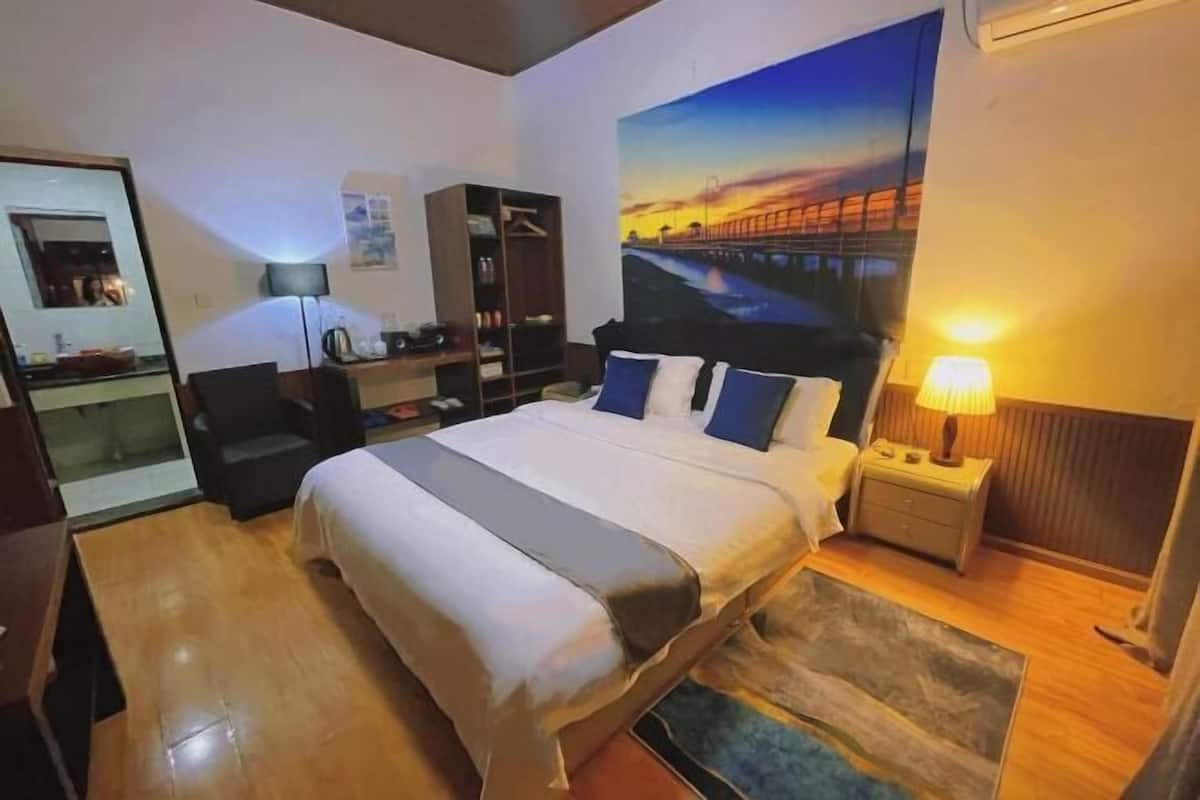 A hotel room with a bed, bedside table, lamp, and a large painting of a sunset on the wall.