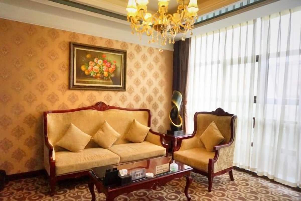 A room with a sofa, armchair, and a glass coffee table. There is a chandelier, a painting on the wall, and a window with curtains.