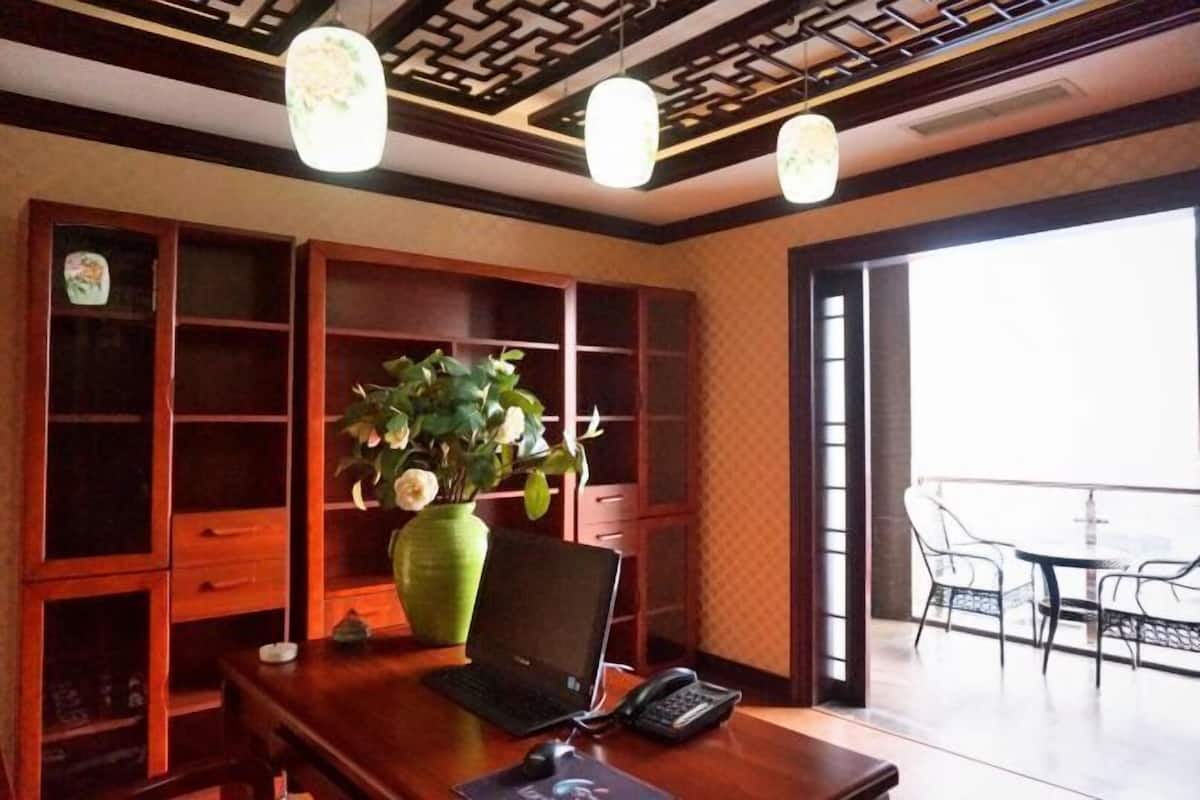 A wooden desk with a laptop, phone, and mouse, a green vase with flowers, and a view of a balcony with chairs.
