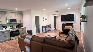 Ein modernes Wohnzimmer mit einer braunen Ledersofa, einem Essbereich mit Tisch und Stühlen, einer Küche mit weißen Schränken und einer Mikrowelle sowie einem an der Wand befestigten Flachbildfernseher.