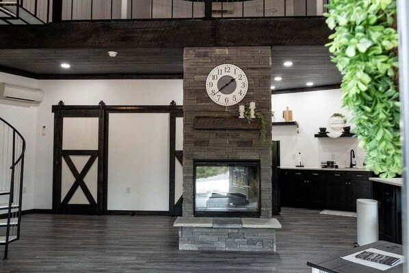 A modern interior with a stone fireplace, a large clock, and a view of the outside through a sliding door. - Grain Bin - Unique Stay (Peebles)