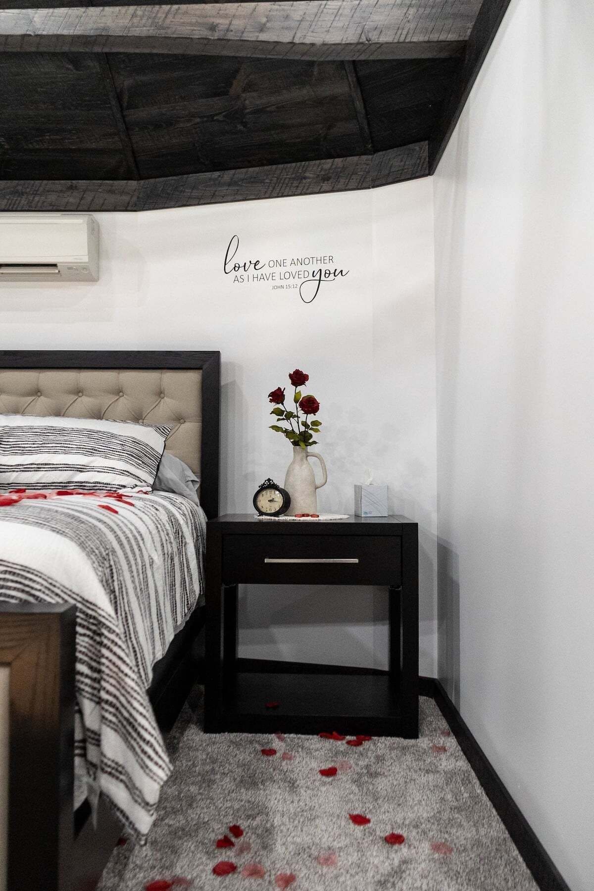 A bedroom with a bed, a nightstand with a vase of flowers, a clock, and a wall with a biblical quote.