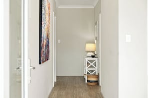 A hallway with a white door, a wall-mounted painting, a white console table with a lamp, and a woven basket.
