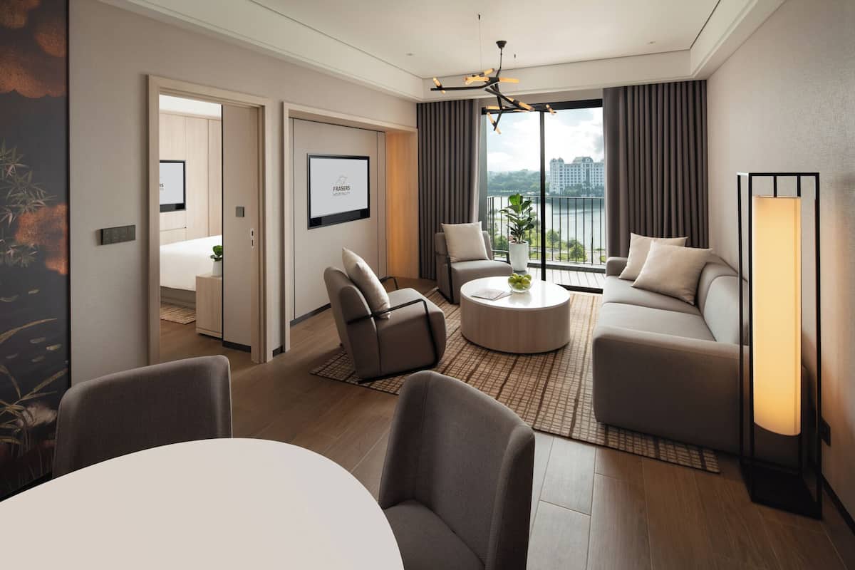 A modern hotel room with a living area, a dining table, and a large window offering a city view.