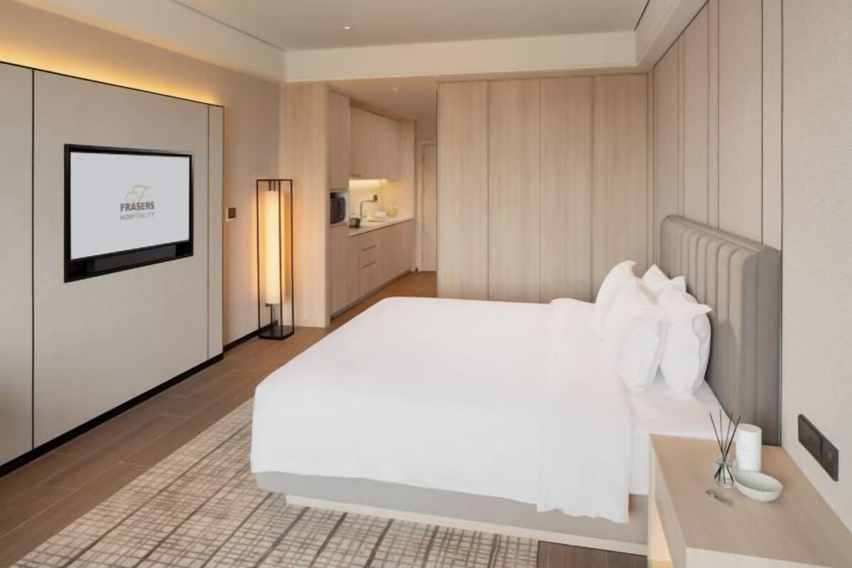 Une chambre d’hôtel avec un grand lit, une table de chevet avec une lampe, un téléviseur fixé au mur et une vue sur le couloir.