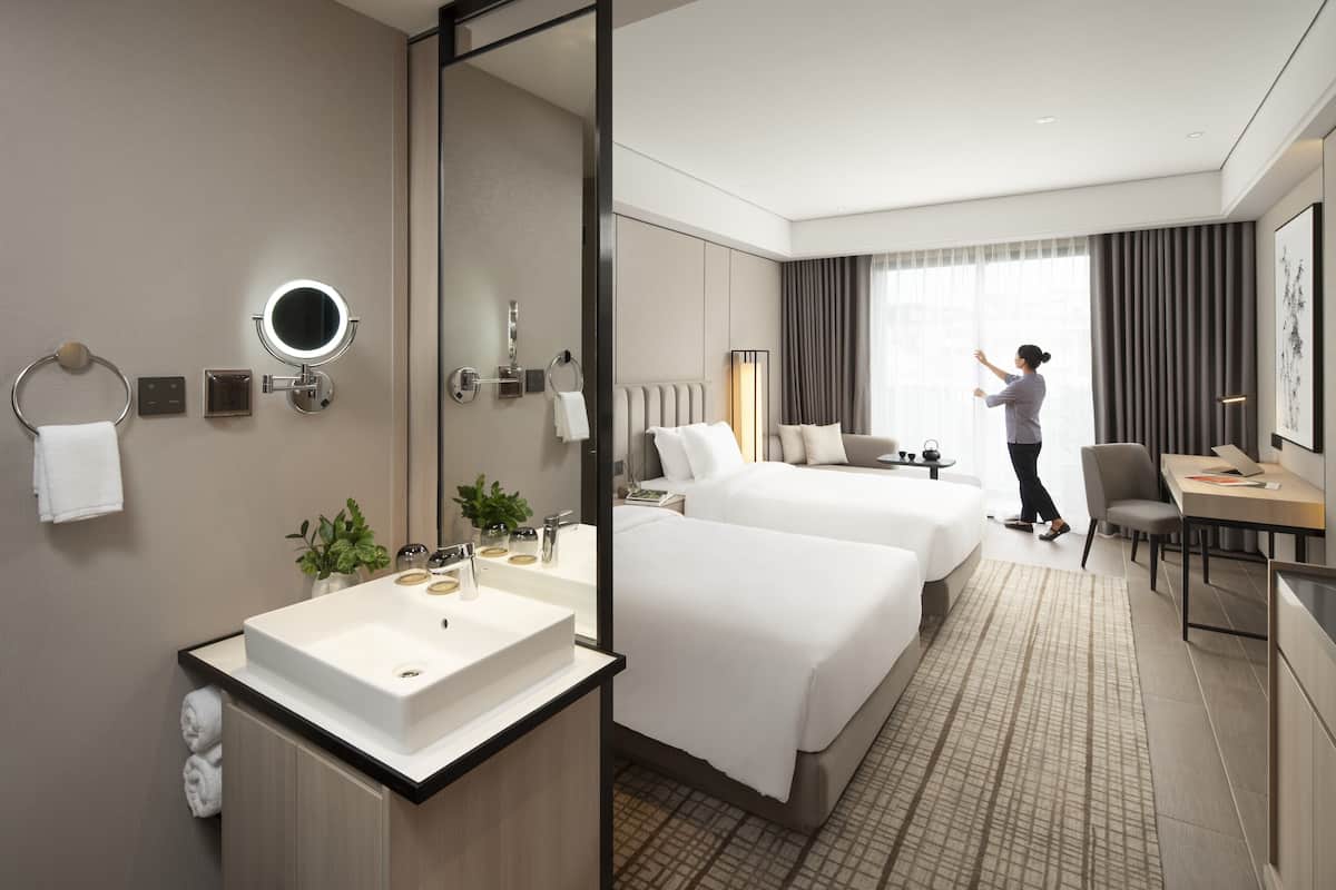 A hotel room with a large bed, a desk, a chair, a bathroom with a sink and mirror, and a large window with curtains.