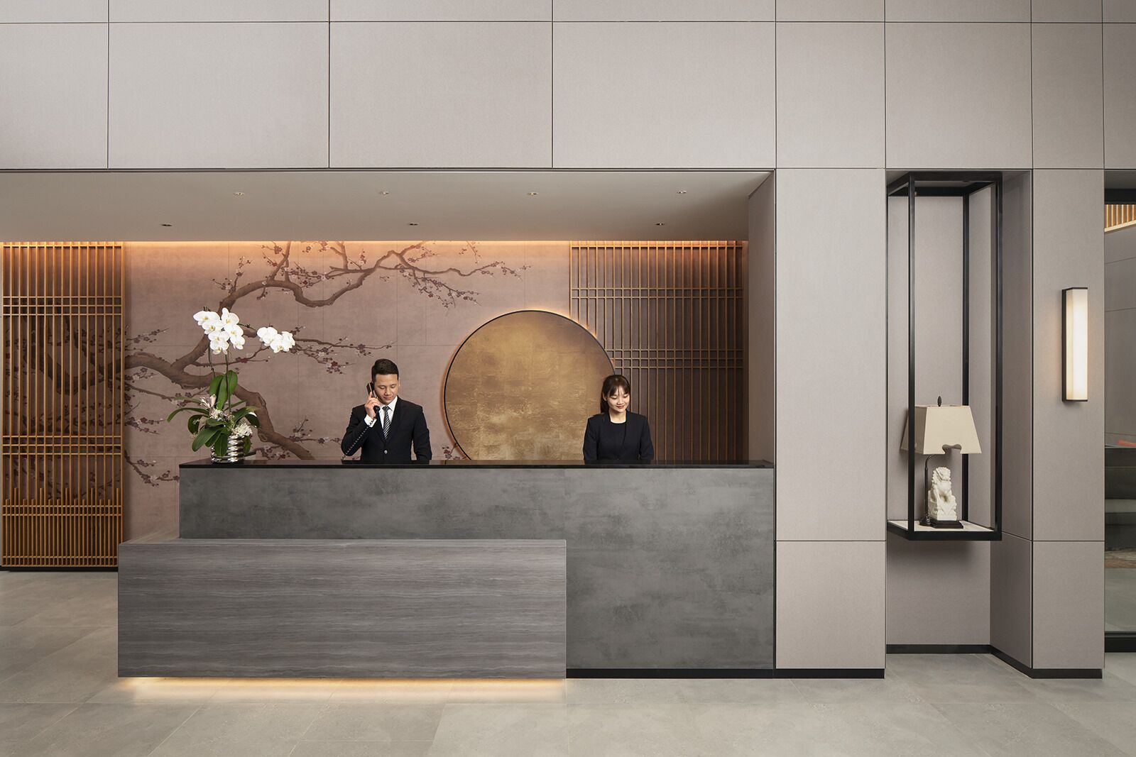 Una moderna recepción con diseño minimalista, que presenta un jarrón grande con flores blancas, un adorno circular grande en la pared y un área de recepción elegante.