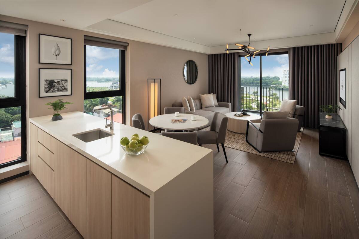 A modern hotel room with a kitchen area, a dining table, a sofa, and a balcony with a view.