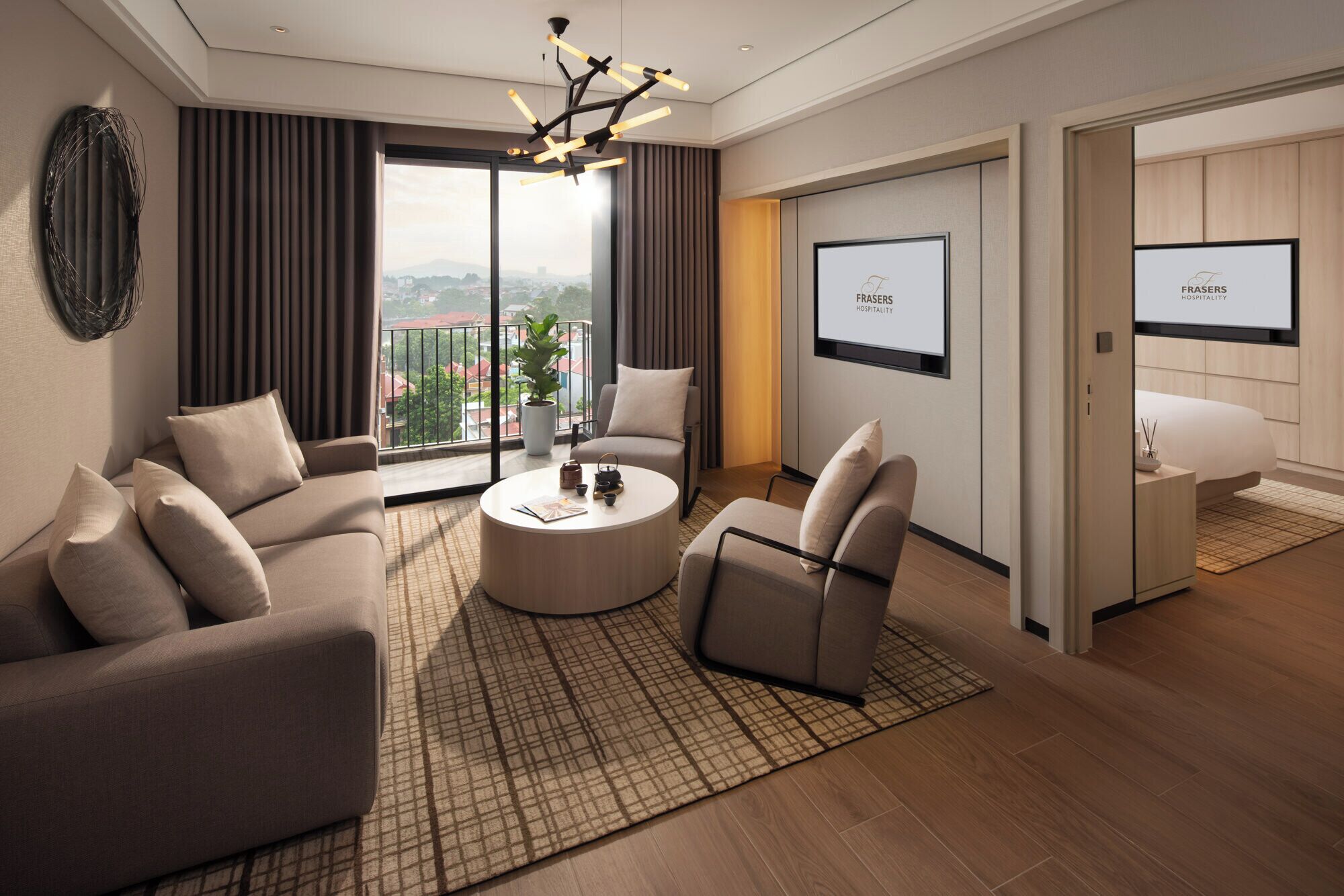 A modern hotel room with a sofa, armchair, and a round coffee table. There is a large window with a view of buildings and a balcony. The room has a flat-screen TV mounted on the wall and a door leading to another room.