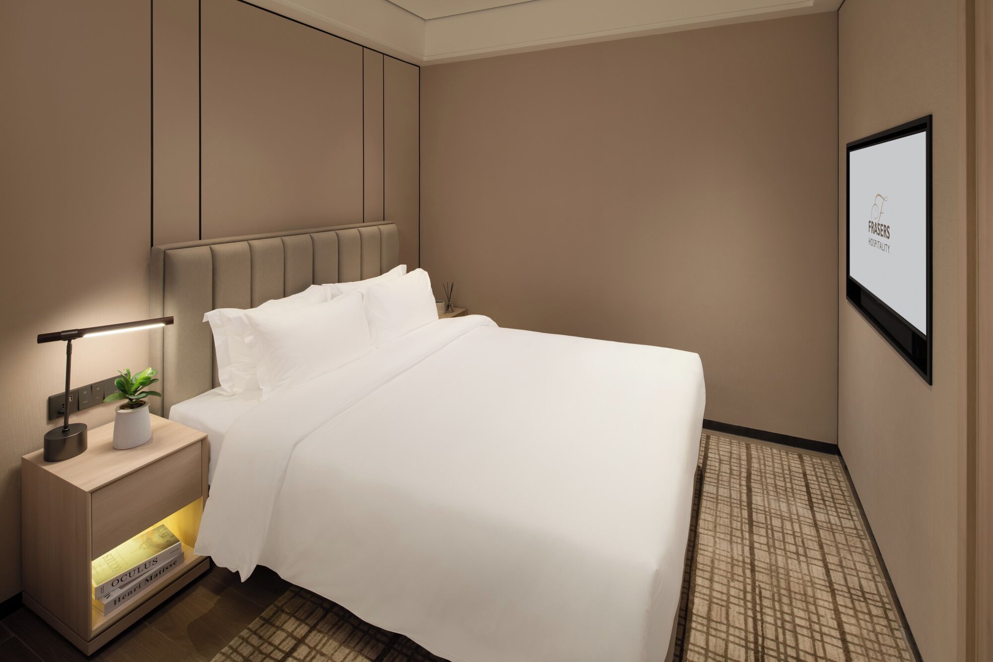 A neatly made bed in a hotel room with a headboard, a bedside table with a lamp, a small plant, and a framed picture on the wall.