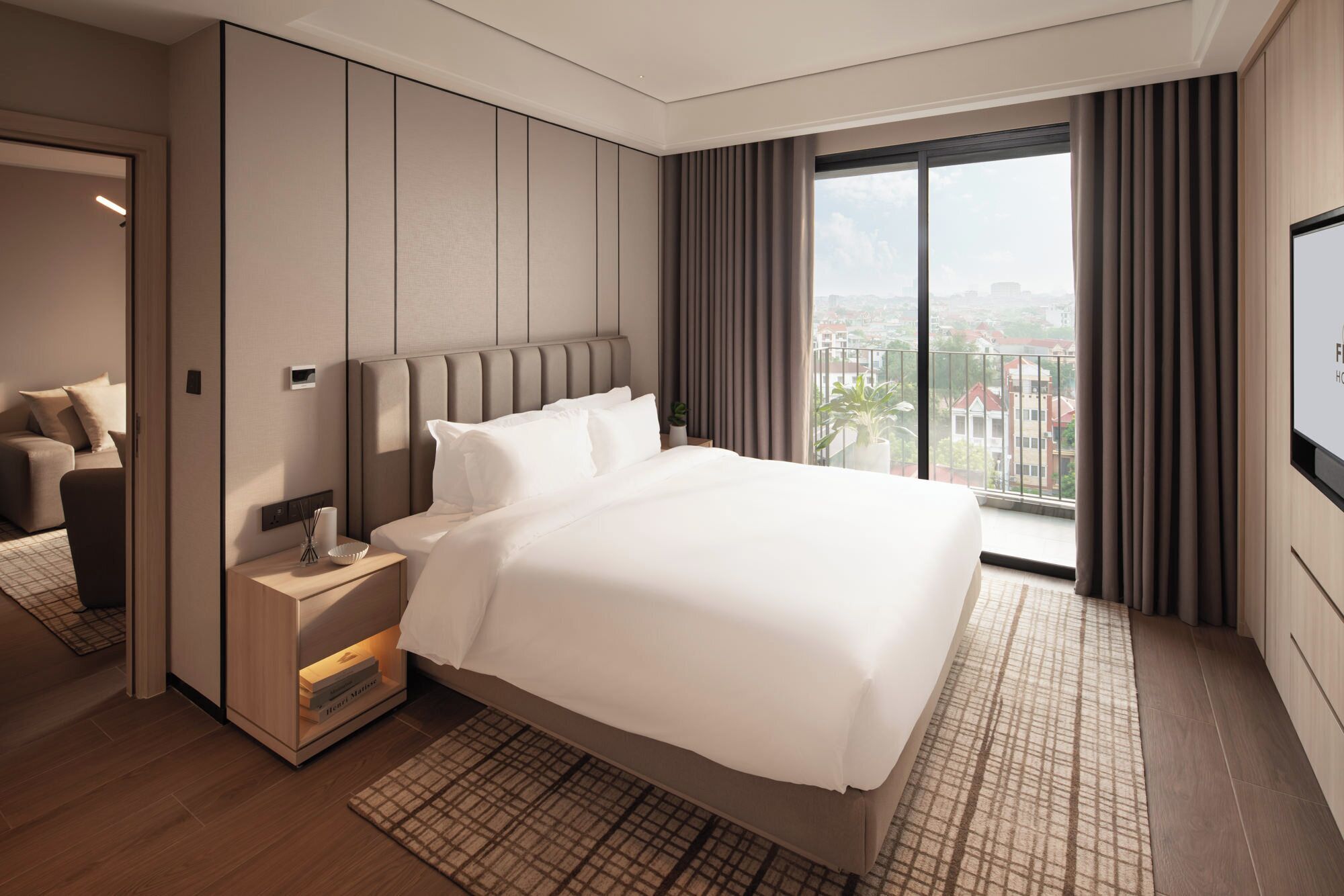 A modern hotel room with a large bed, a bedside table, a TV, and a view of the city.