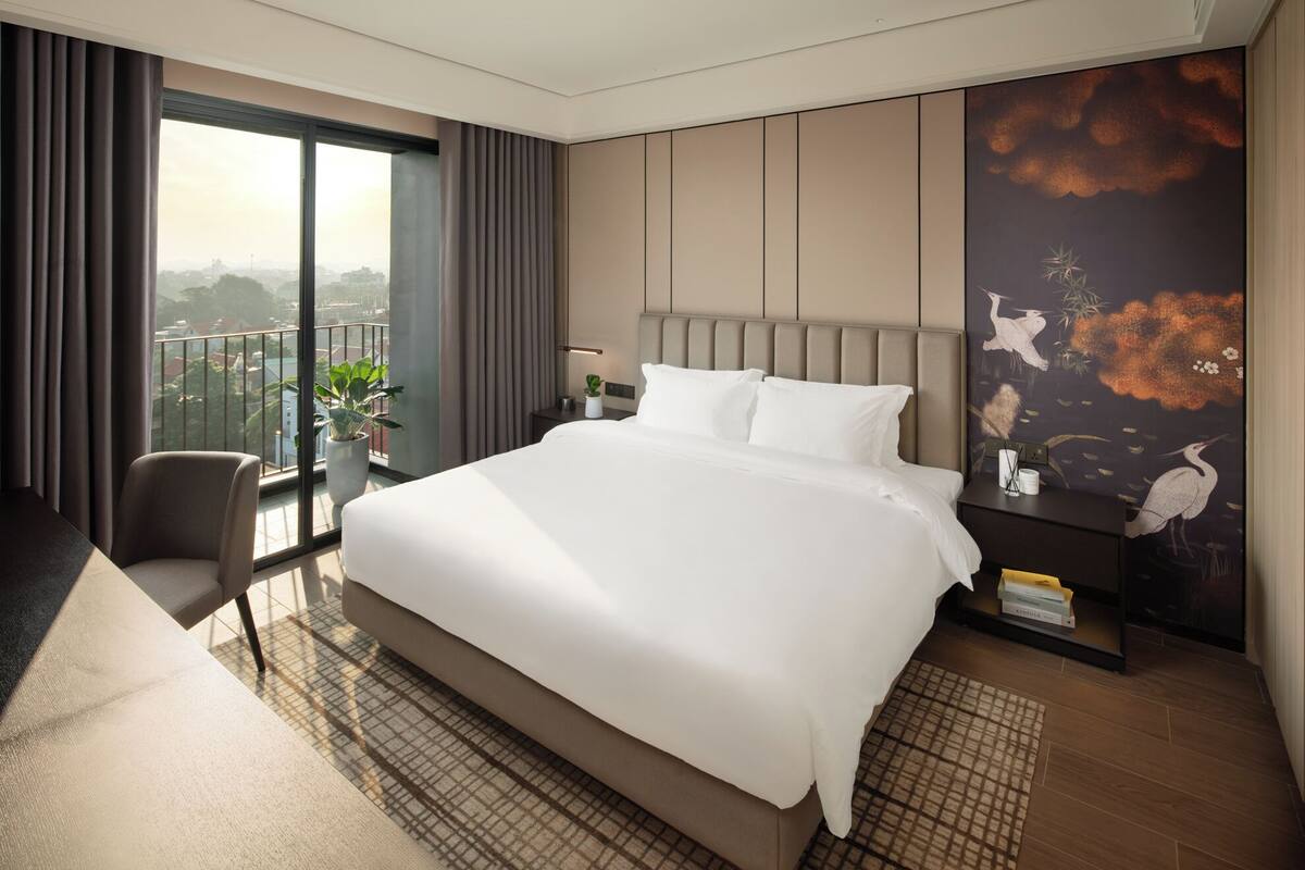 A modern hotel room with a large bed, a balcony with curtains, a desk, and a wall with a nature-themed mural.