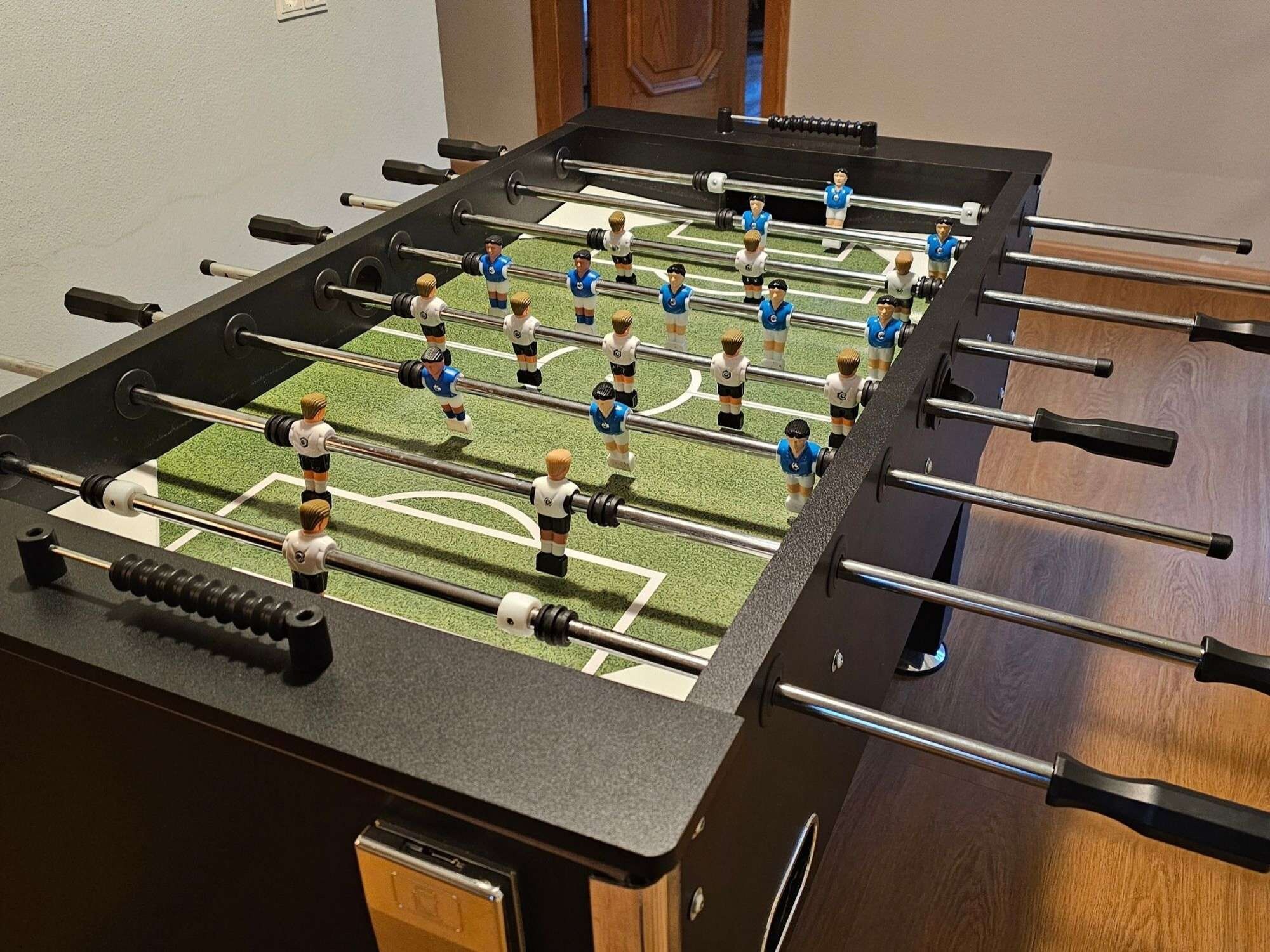 Ein Kicker mit dunkler Spielfläche und mehreren Stangen, mit Spielerfiguren in blauen und weißen Trikots.