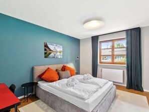 A hotel room with a bed, pillows, a painting, a window with curtains, and a red table.