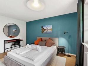 A modern bedroom with a bed, a desk, a chair, a round mirror, a painting, and a floor lamp.