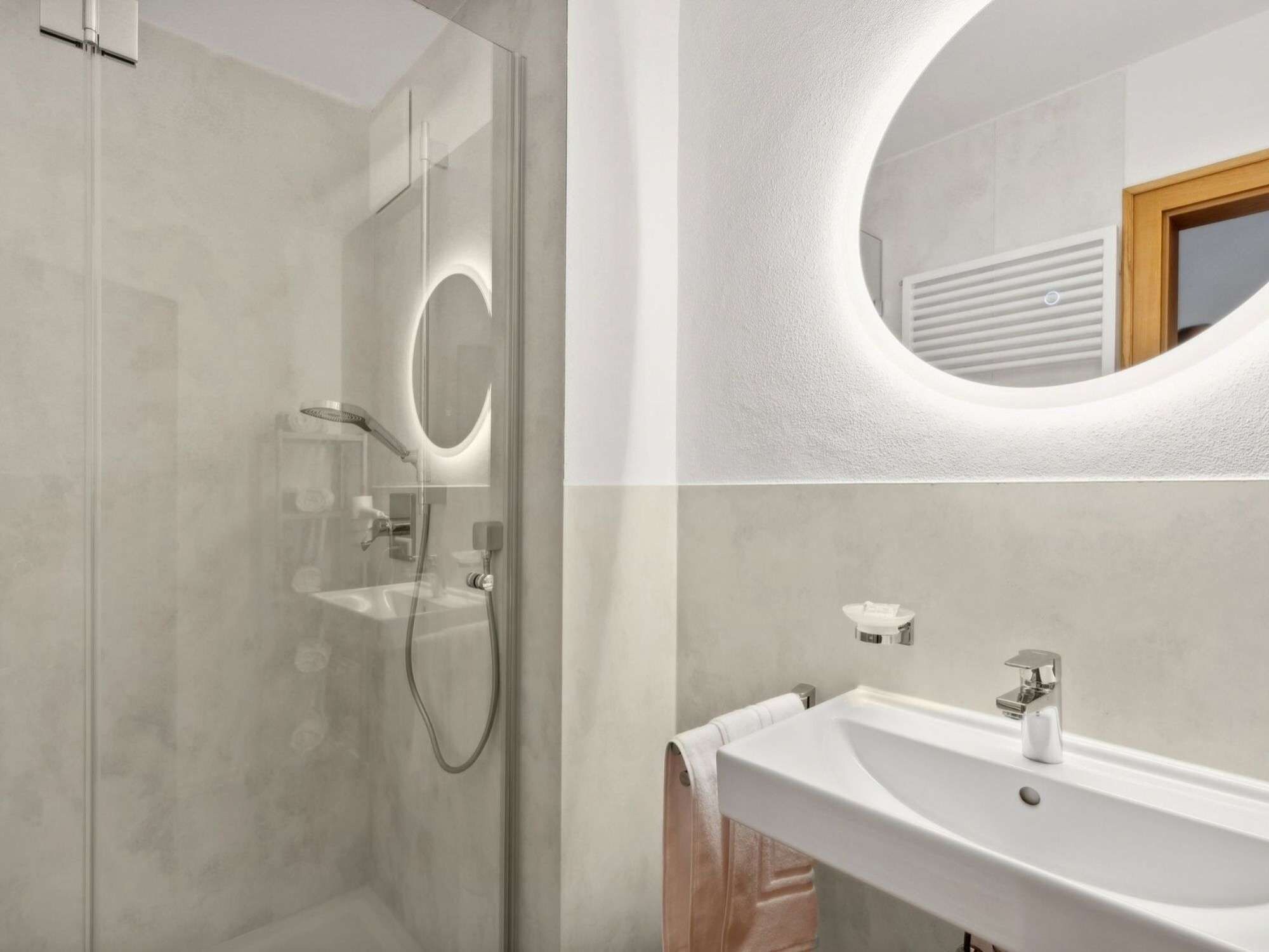 A modern bathroom with a glass-enclosed shower, a wall-mounted sink, and a round mirror with a light.