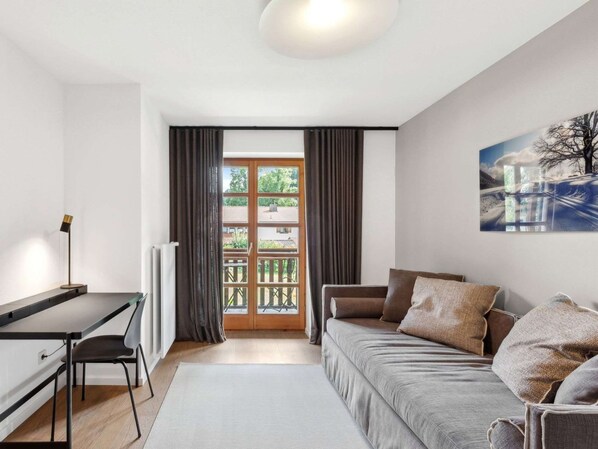 A modern living room with a sofa, a desk, and a chair. There is a large window with curtains, a radiator, and a painting on the wall.
