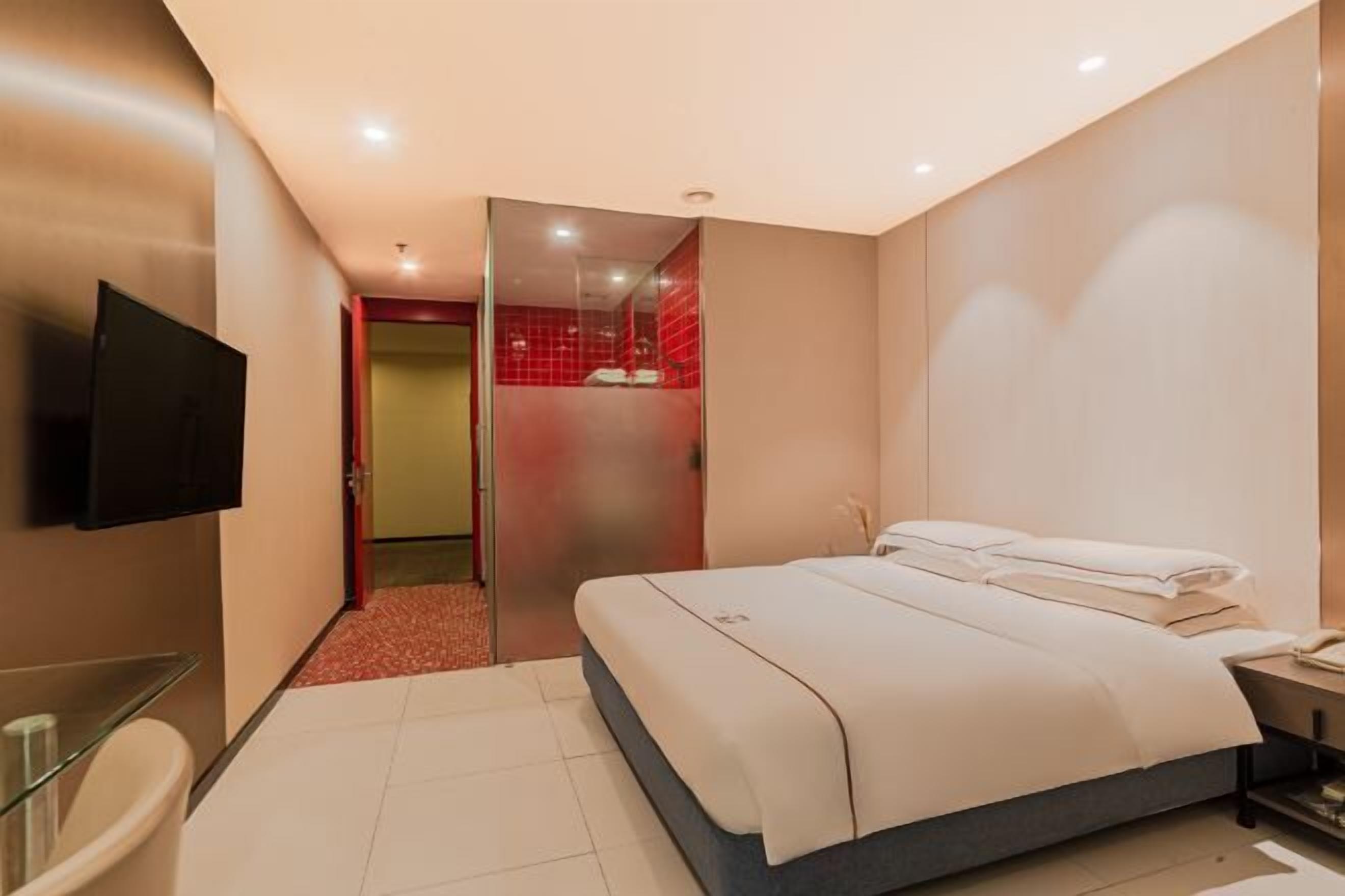 A modern hotel room with a bed, a TV, a glass shower enclosure, and a red-tiled bathroom.