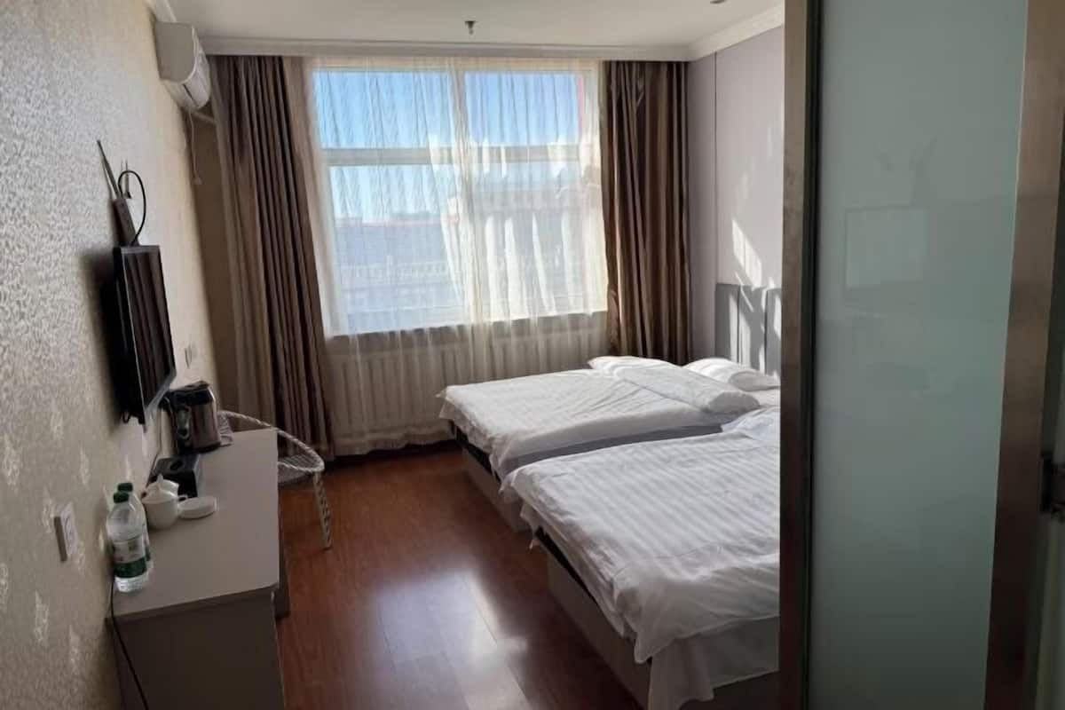 Habitación de hotel con dos camas, un escritorio pequeño con una silla, un radiador y una ventana con cortinas.