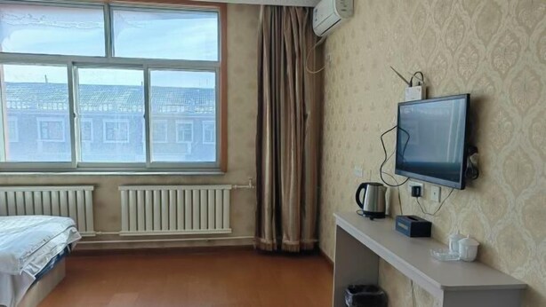 A hotel room with a bed, a TV, a radiator, a window with curtains, and a view of buildings outside. - Haojin Business Hotel (Erenhot)