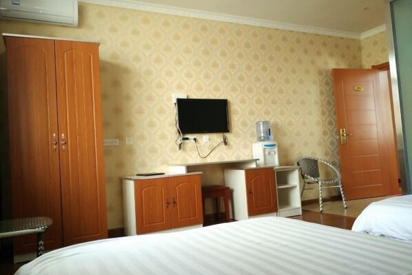 A hotel room with a bed, wooden wardrobe, desk with a chair, a television, and a water dispenser.